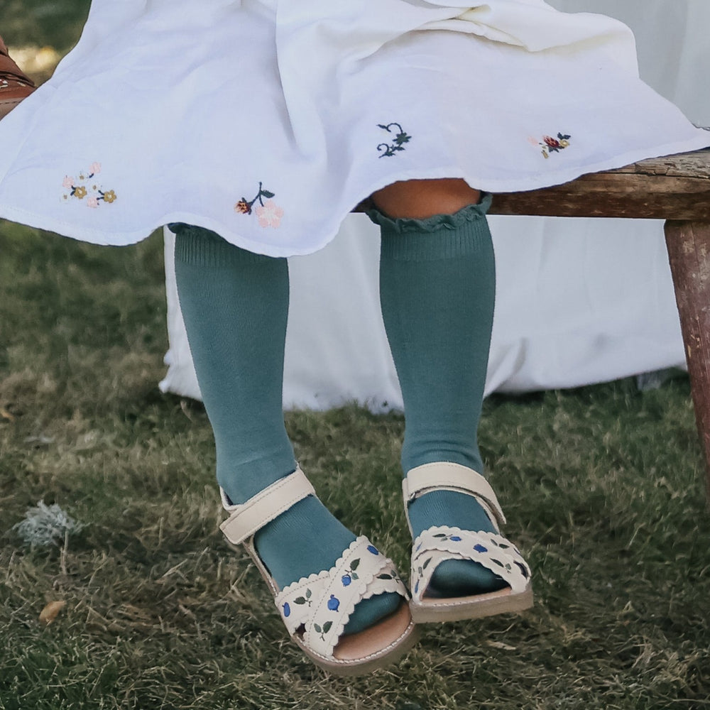 Photo des sandales brodées blueberry crème de la collection Uniqua de chez Petit Nord portées par une petite fille assise sur un banc