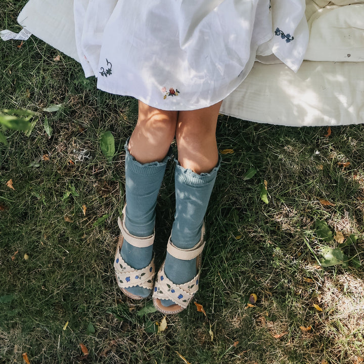Photo des sandales brodées blueberry crème de la collection Uniqua de chez Petit Nord portées par une petite fille allongée dans l'herbe
