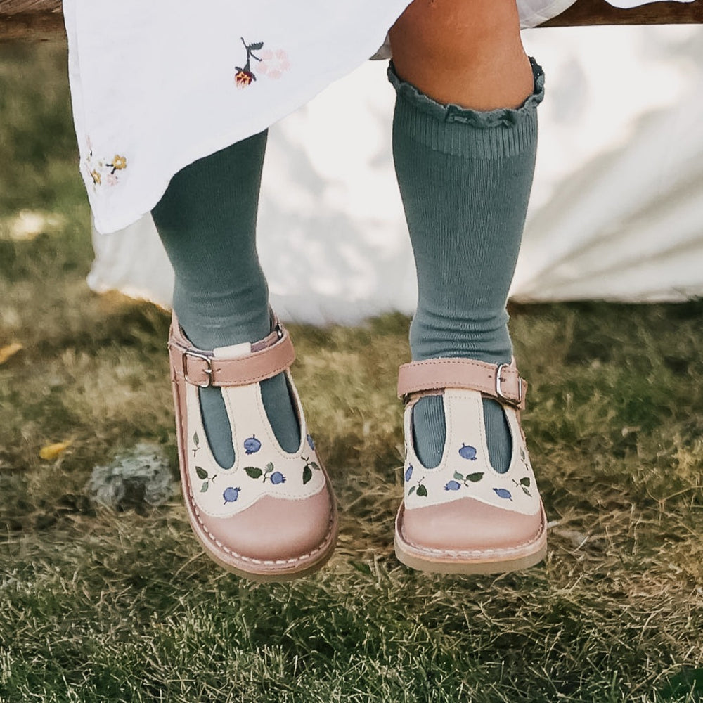 Photo des babies Peter Pan brodées rose de la collection Uniqua de chez Petit Nord portées par une petite fille assise sur un banc en bois